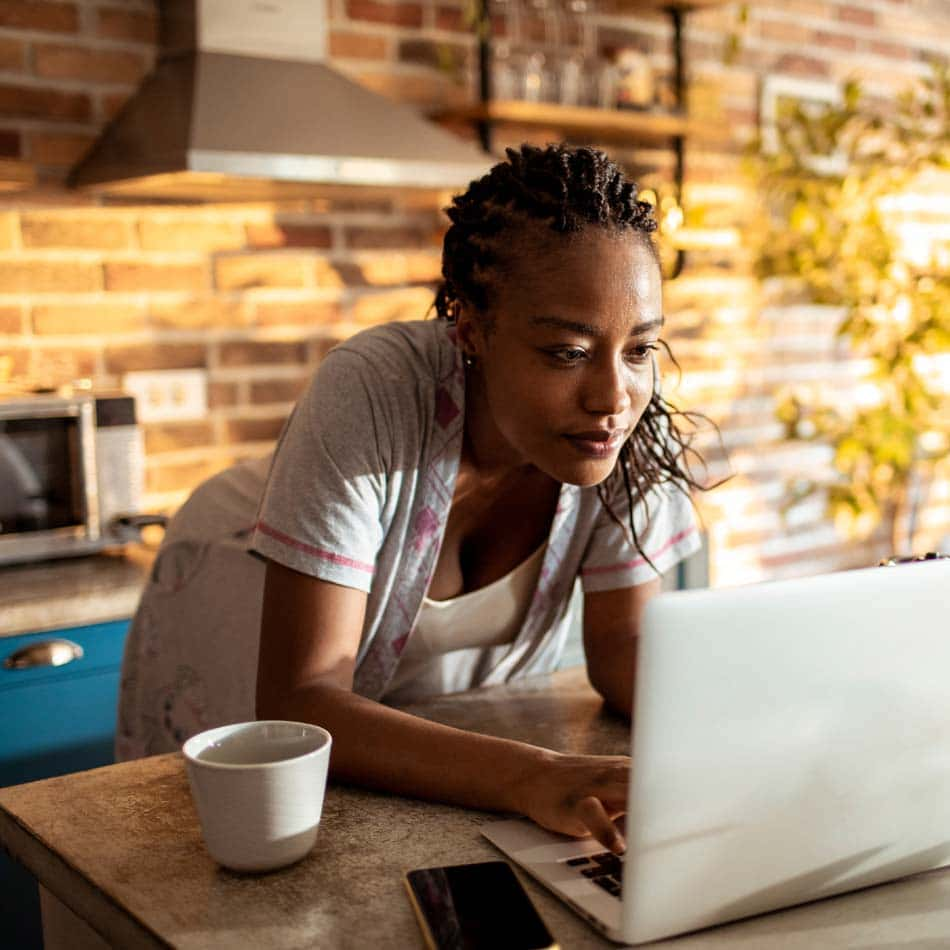 Woman in her kitche looking at her laptop. She has a cup of coffee and is smiling.